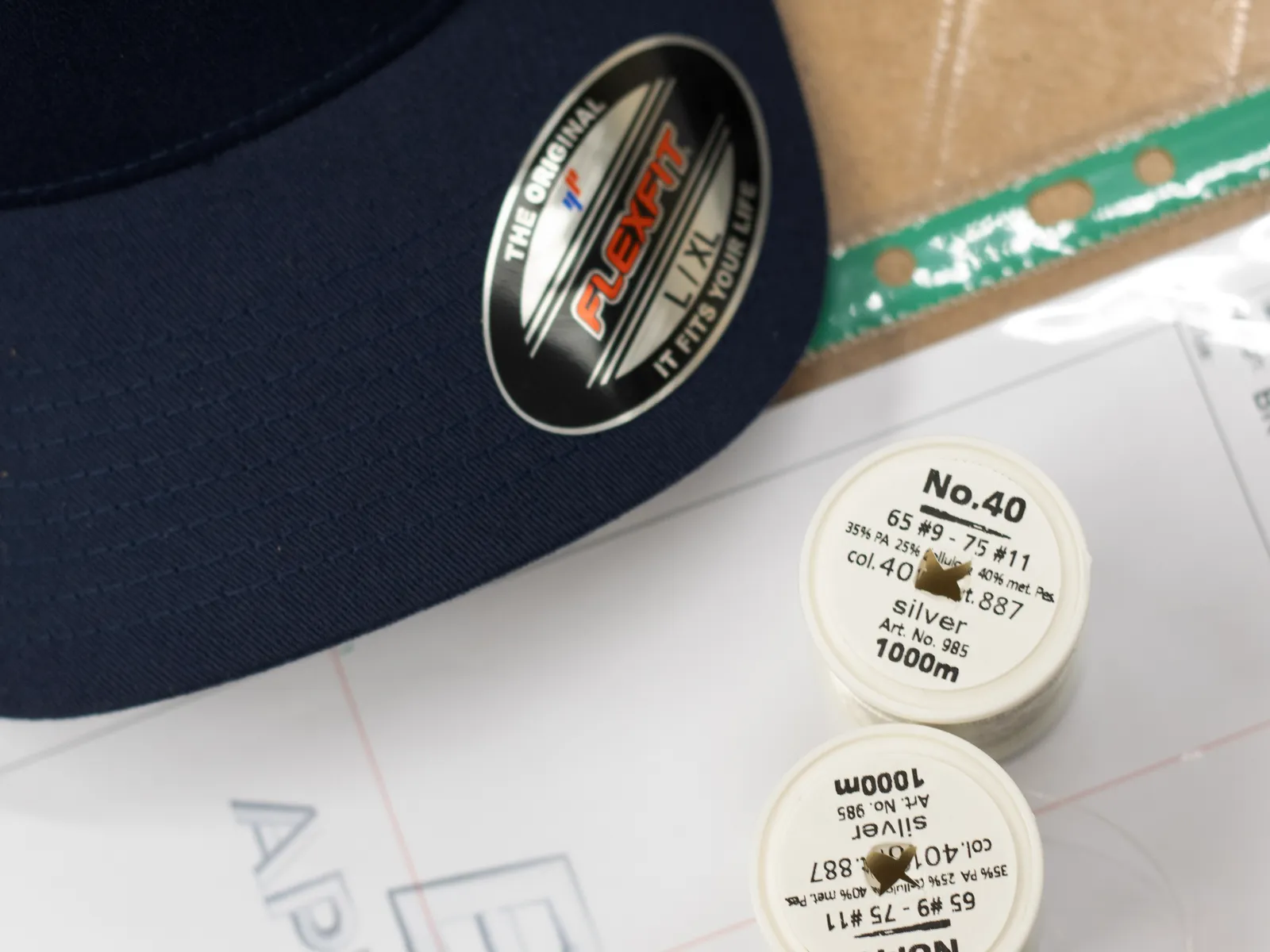 A navy Flexfit cap beside spools of silver thread and the digitising sheet for the logo